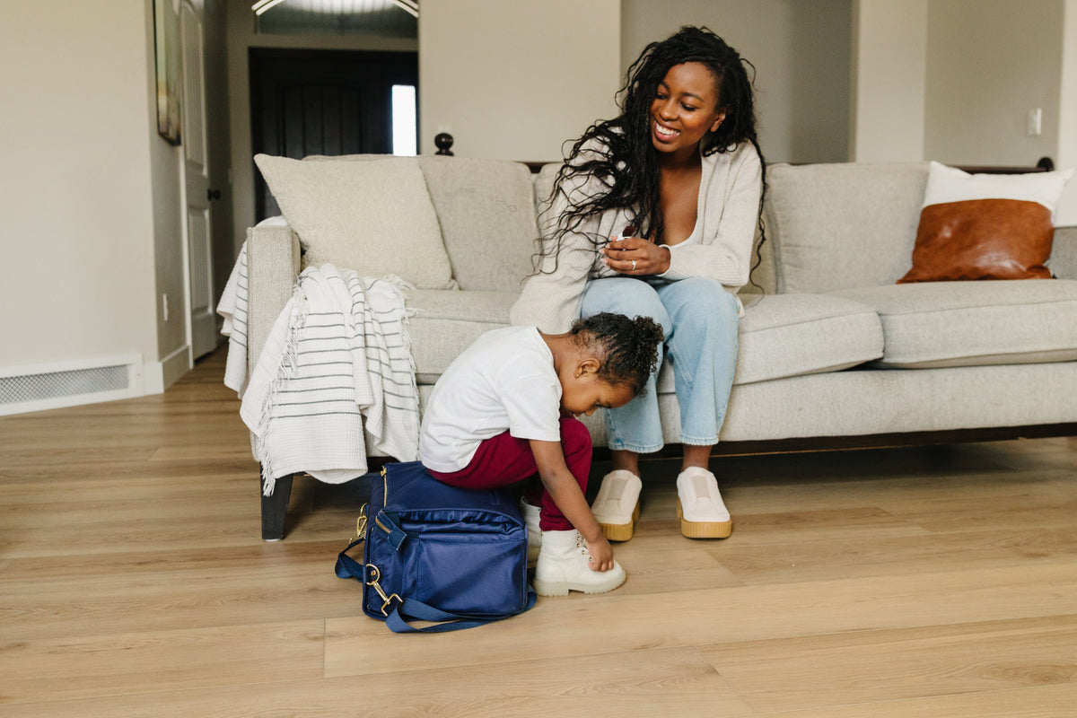 Mother watching child get ready beside the August & Ivy IvyGo diaper bag, showing real-life everyday parenting convenience.